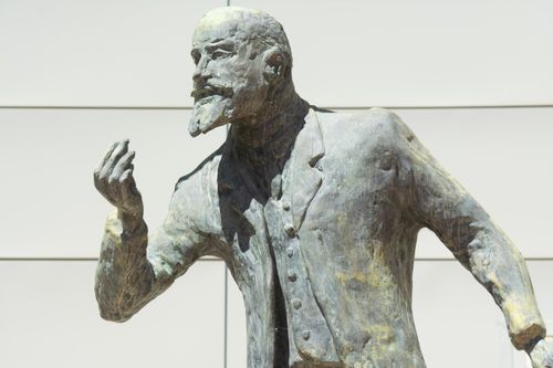 Free Bronze sculpture of a historical figure in an assertive pose, captured outdoors against a modern building. Stock Photo