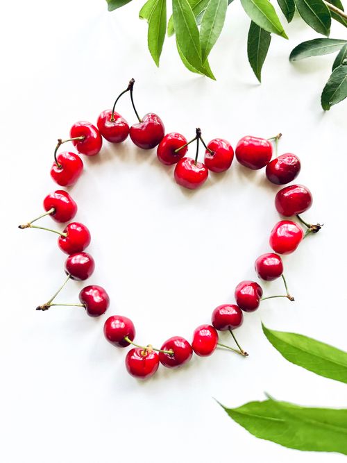 Free A creative heart-shaped cherry arrangement surrounded by green leaves on a white background. Stock Photo
