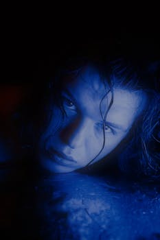 Close-up of a woman's face with blue lighting and wet hair on a dark background.