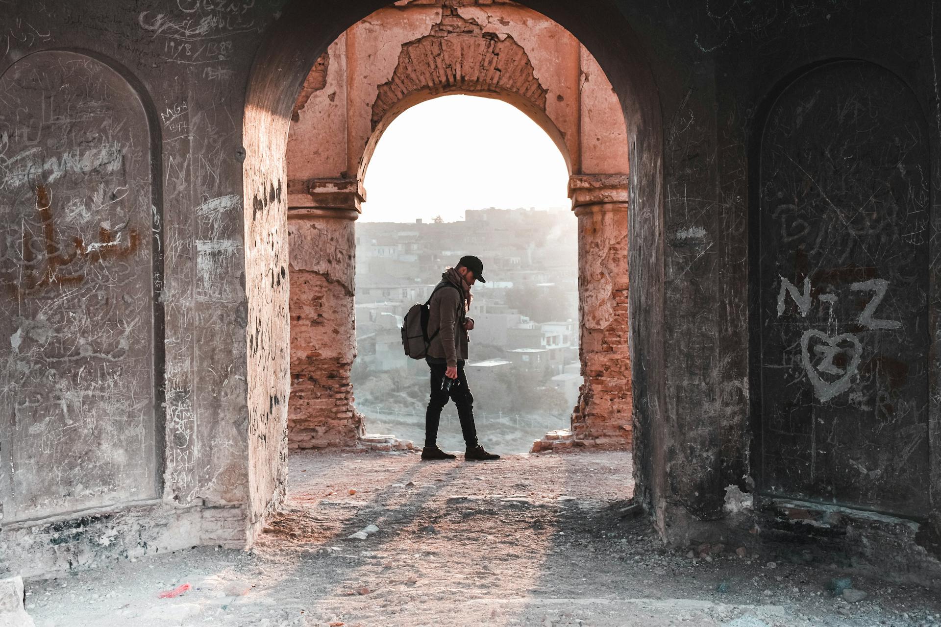 Silhouette of a traveler with a backpack walking through a historic stone archway at sunset.