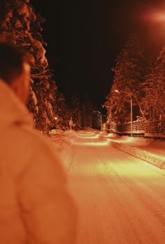 A person stands on a snowy road illuminated by streetlights in a winter nighttime setting.