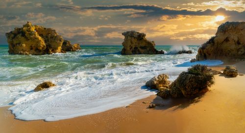 Free Scenic view of a rocky seashore at sunset with waves crashing under a golden sky. Stock Photo