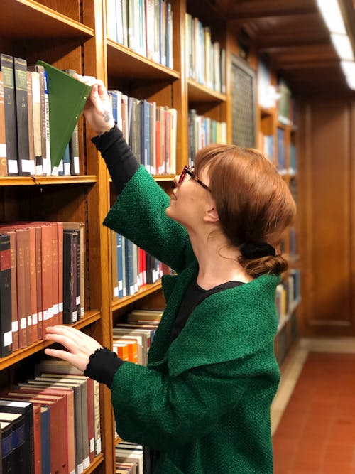 Woman Wearing a Green Blazer Taking a Book from a Wooden Bookcase