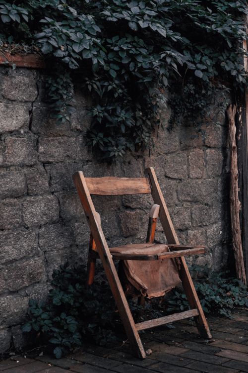 Free A worn wooden chair leaning against a stone wall surrounded by leaves. Stock Photo