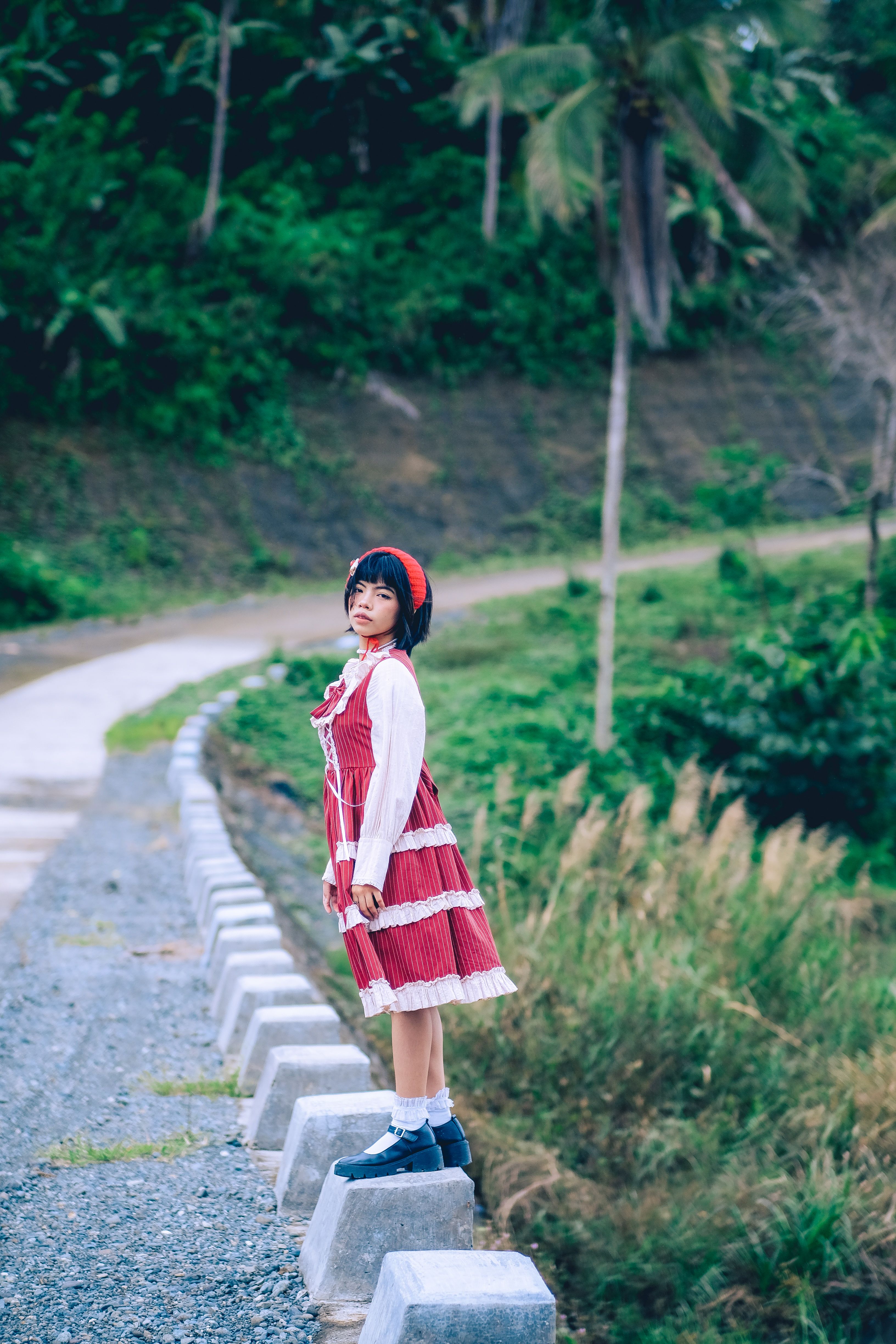 A young girl in a red dress standing on a roadside with lush greenery in the background, creating a serene landscape.
