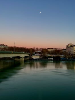 Peaceful twilight view of a cityscape with a river, bridge, and moon visible.