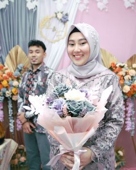 Portrait of a smiling bride in traditional attire holding a bouquet, captured during a colorful wedding ceremony. - Fotografer Sidoarjo