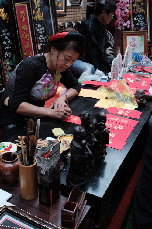 Free Vietnamese woman writing calligraphy in a vibrant souvenir shop with cultural items displayed. Stock Photo