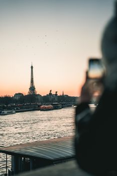 A stunning sunset view of the Eiffel Tower captured from the Seine River with a person taking a photo in the foreground.
