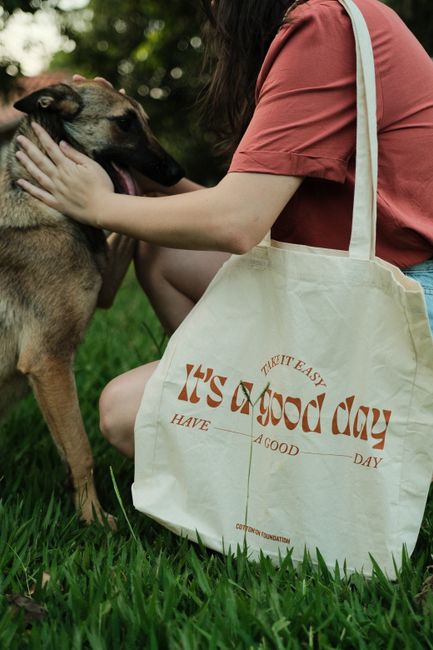 A woman in a park petting a dog with a tote bag that reads 'It's a good day'.