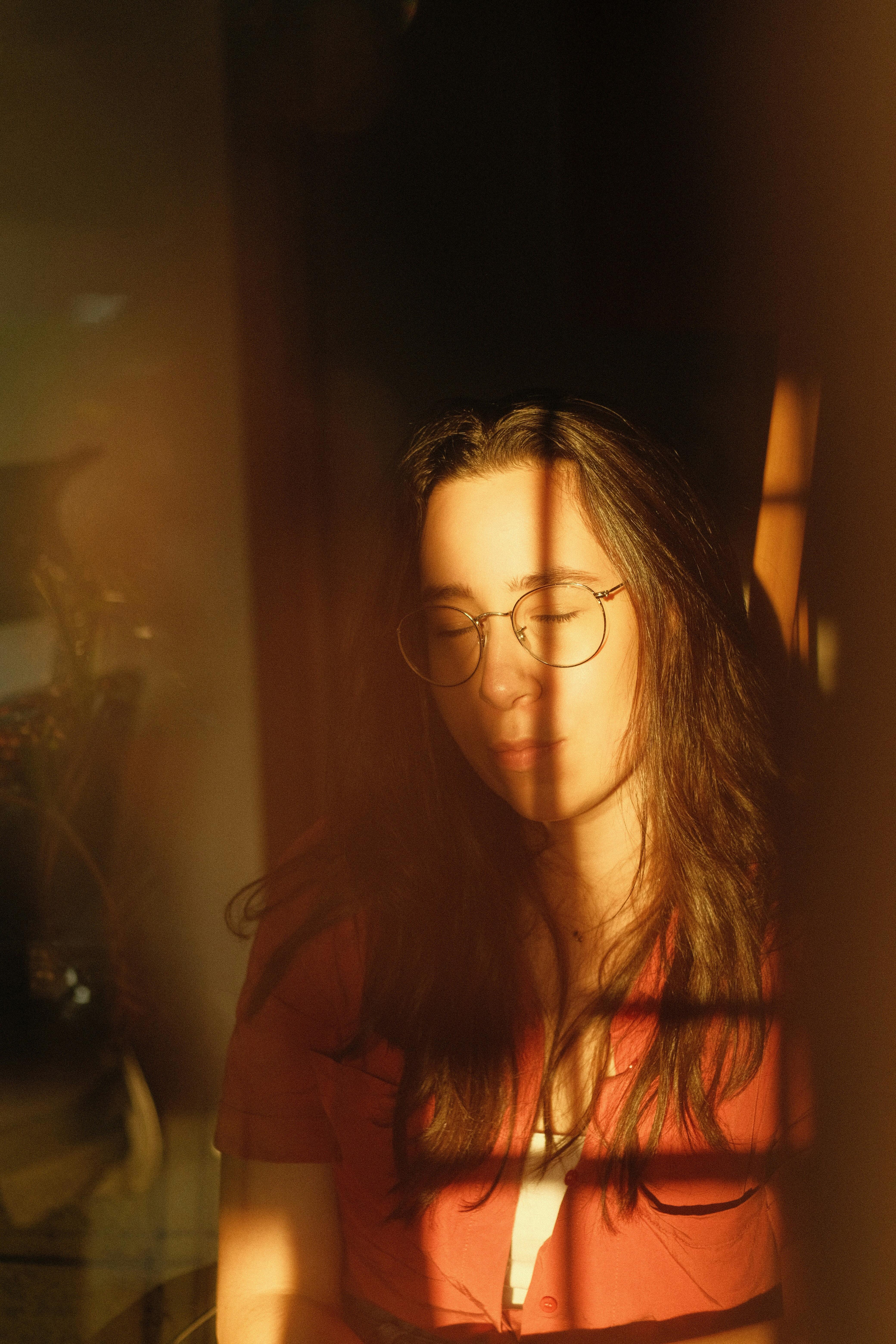 Peaceful woman with glasses enjoying warm sunlight indoors, eyes closed.