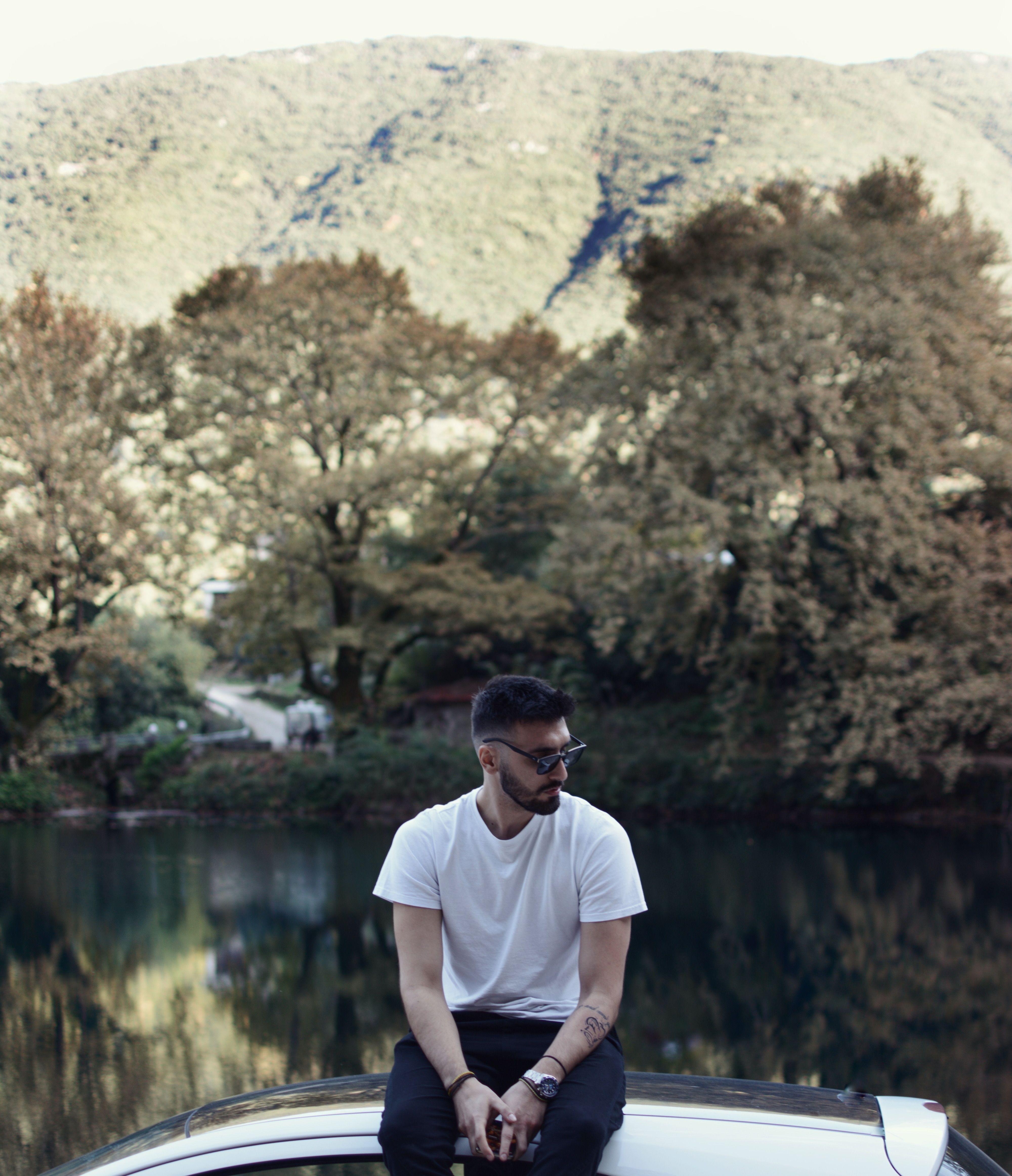 A man in sunglasses sits on a car roof by a tranquil lake with a mountainous backdrop.