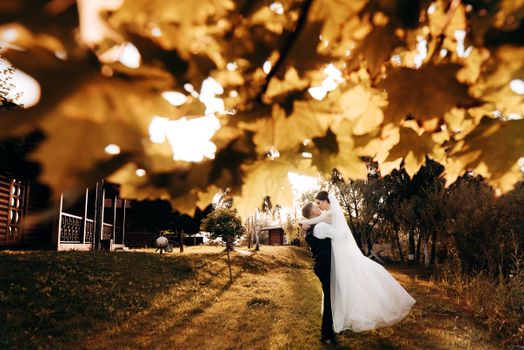 Bride and groom share a kiss under vibrant autumn leaves during sunset in a park setting.