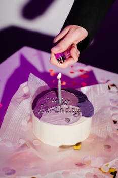 Hand lighting candle on a purple birthday cake surrounded by confetti.