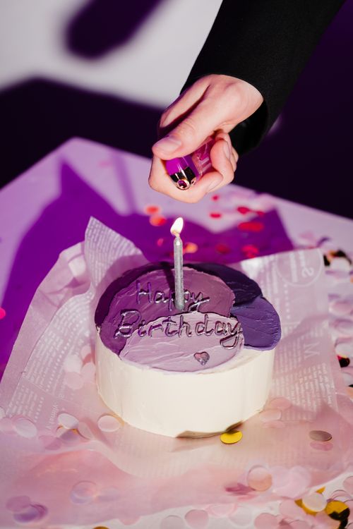 Free Hand lighting candle on a purple birthday cake surrounded by confetti. Stock Photo
