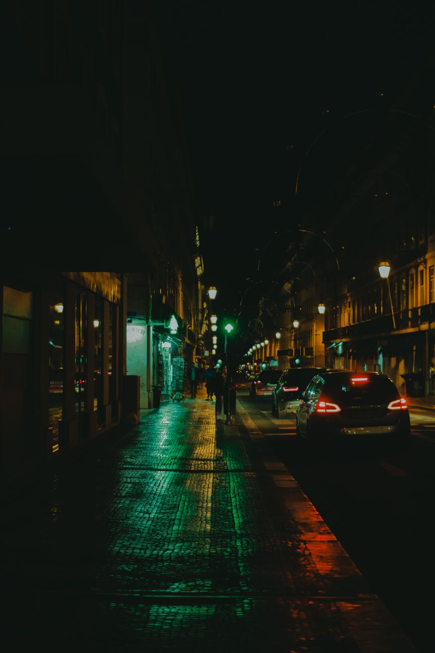 Street view with cars and people, illuminated by streetlights on a wet road at night.