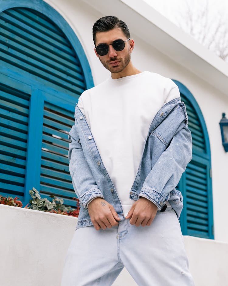 Man Wearing Jean Jacket And Sunglasses Standing By Wall With Blue Window Shutters