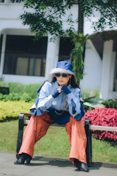 Stylish woman in a hat poses outdoors in a sunny Bandung garden, showcasing vibrant fashion.
