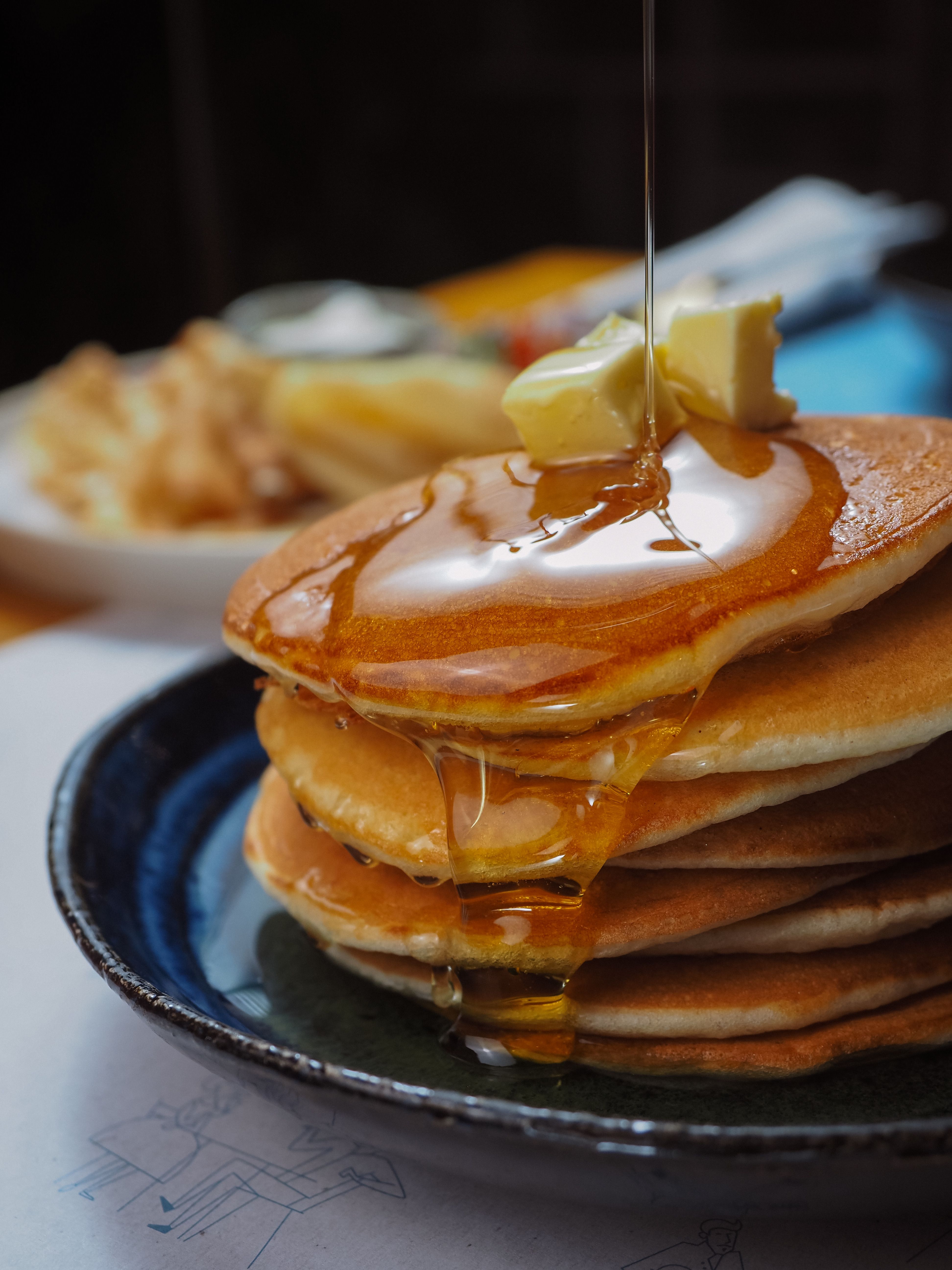 A tall stack of golden hotcakes on a plate with a pat of melting butter on top and syrup dripping down the sides.