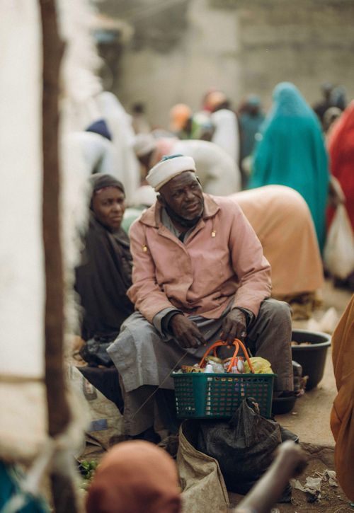 Free Elderly man sitting with shopping basket in bustling market, surrounded by people. Stock Photo