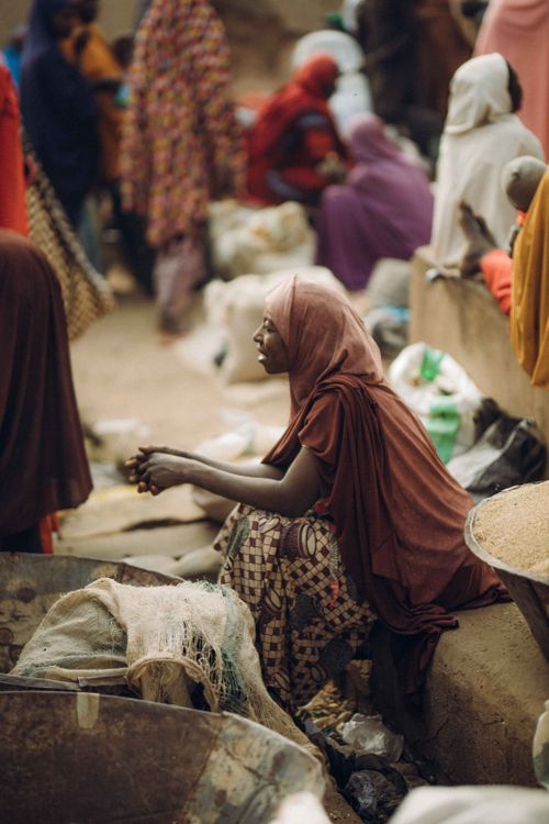 Free A woman in traditional clothing sits at an outdoor market, surrounded by people and goods. Stock Photo
