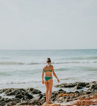 A woman in a green bikini walks along a rocky ocean shore, enjoying a sunny day.