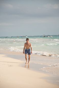 Shirtless man walks on a sandy beach by the sea, enjoying summer vibes.