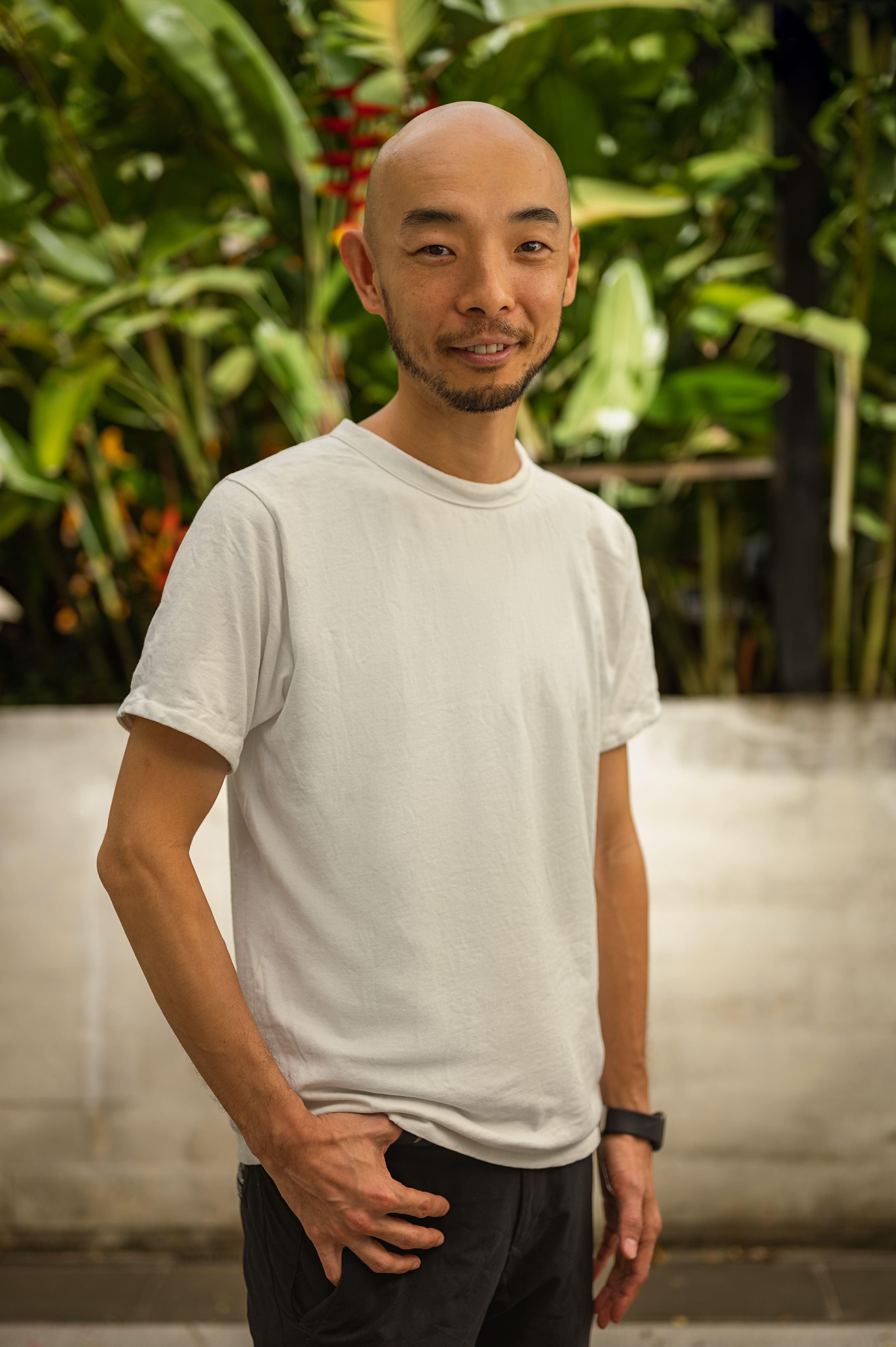 Bald Asian man in a white t-shirt posing outdoors with lush greenery.