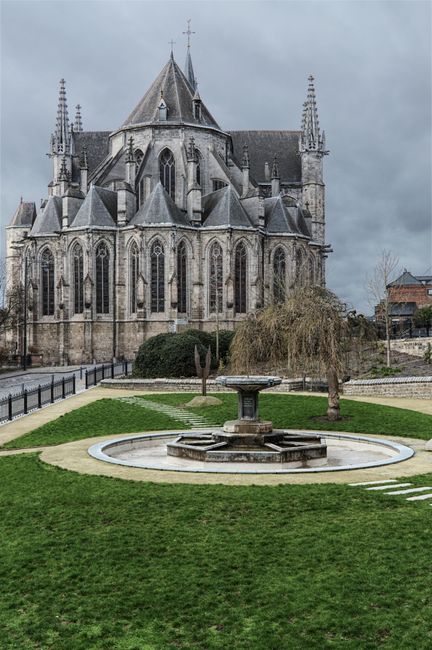 Gothic cathedral in Mons, Belgium with stone fountain and green garden in overcast weather.
