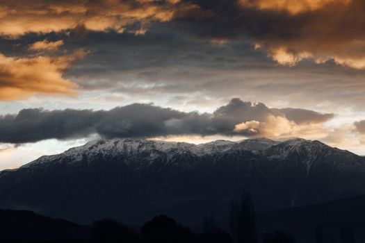 Snowcapped mountains under a dramatic stormy sky during sunset, showcasing nature's grandeur.
