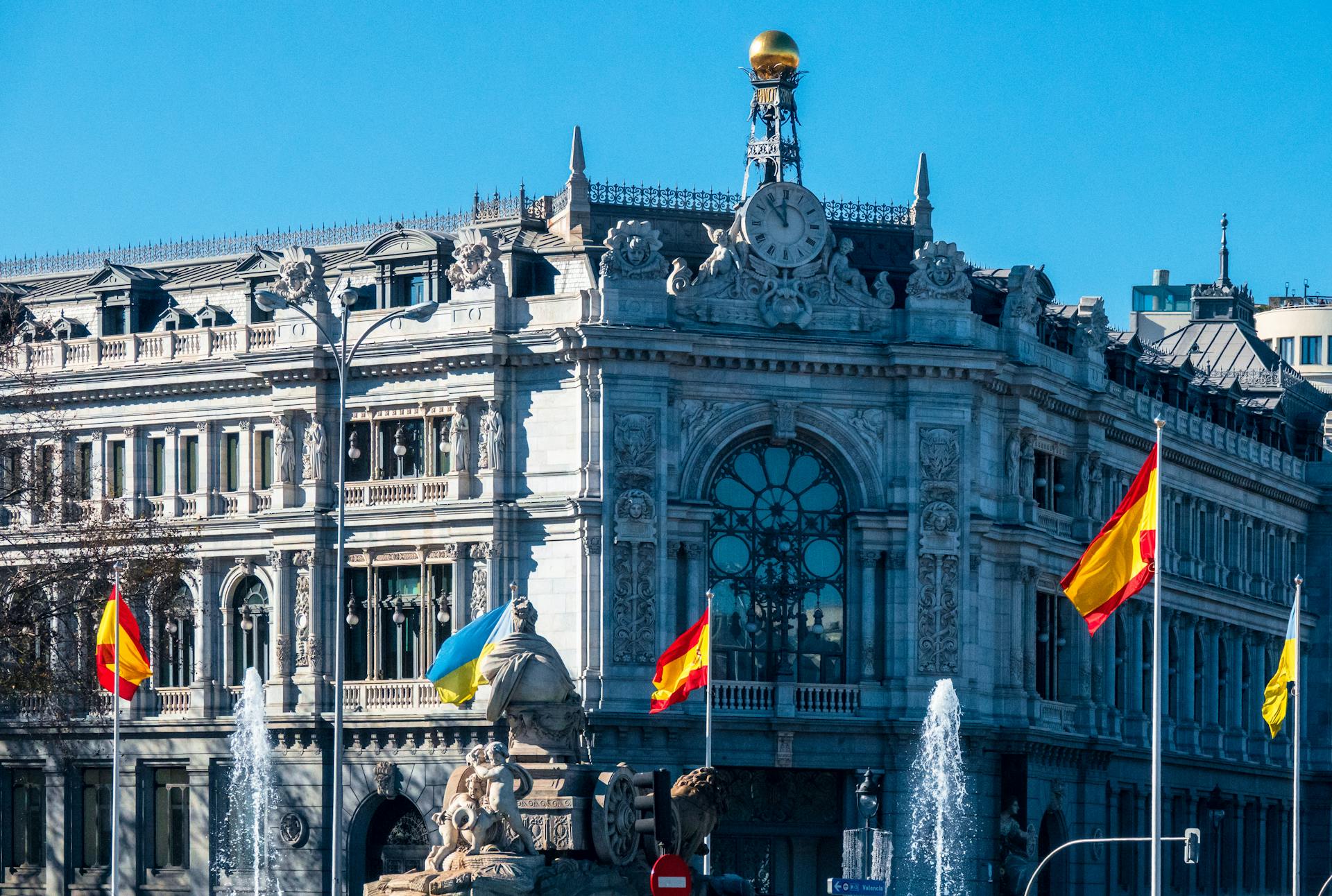 View of the iconic Bank of Spain building and Fountain of Cybele, adorned with flags in Madrid.