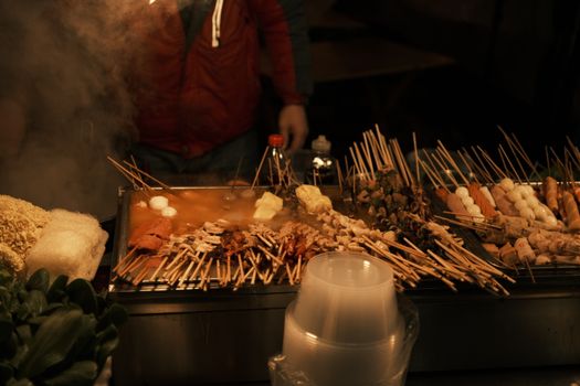 A vibrant display of various skewered meats cooking at a bustling night market food stall.