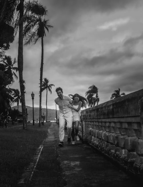 Woman and Man Running Together on the Road by the Beach Under a Cloudy Sky in Black and White