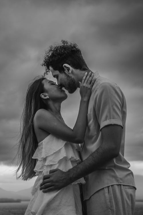 Young Woman and Man Holding Each Other and Kissing in Front of a Cloudy Sky in Black and White