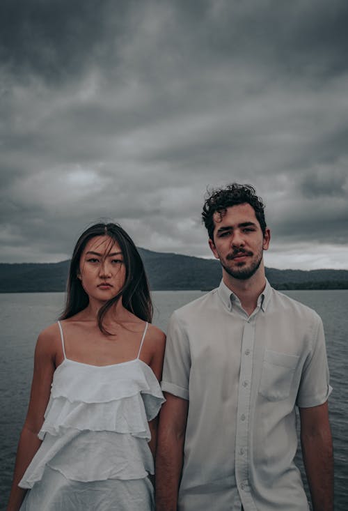 Young Woman and Man Standing Next to Each Other in Front of the Sea, Mountains and Under a Cloudy Sky
