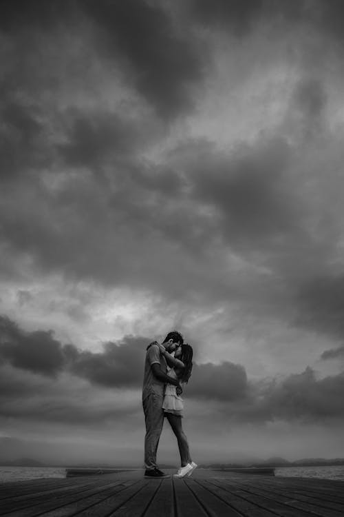 Woman and Man Hugging Each Other on the Pier Under Cloudy Sky in Black and White