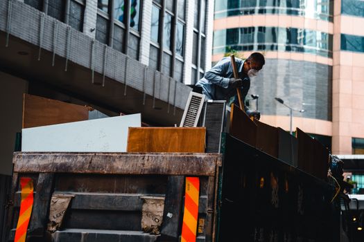 A man wearing a mask manages construction debris in a city environment from a large truck.
