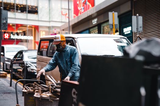 A masked worker collects scrap metal from bins on a city street.