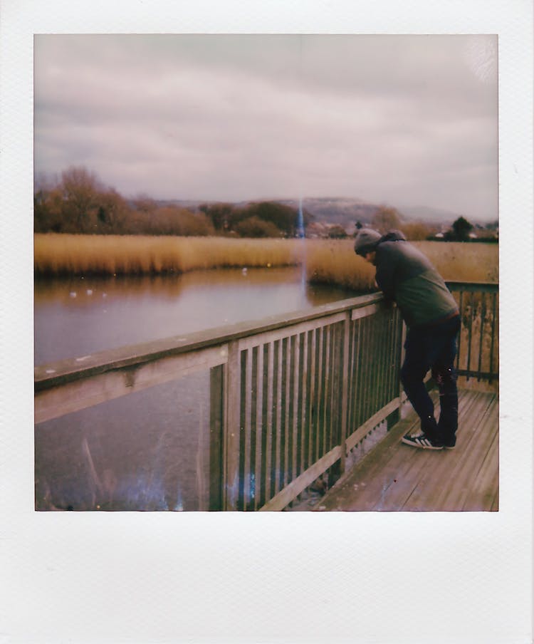 Polaroid Of A Man Leaning Against A Banister And Looking At Water