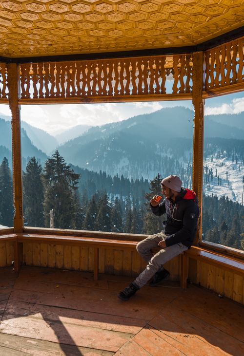 Free Man sitting in a wooden gazebo overlooking snow-covered mountains in Doodpathri, drinking coffee. Stock Photo