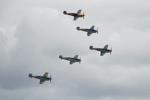 Five historic World War II fighter planes flying in formation beneath a cloudy sky.