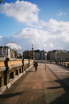 A man cycling along a city bridge with a Ferris wheel and buildings in the background under a blue sky.