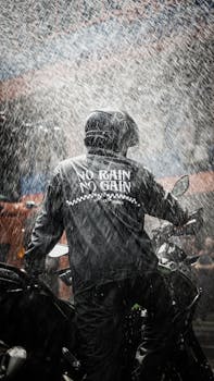 Back view of a motorcyclist riding in heavy rain, wearing a helmet and a jacket with 'No Rain No Gain' text.