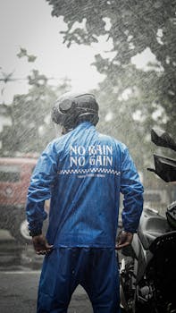 A person standing outdoors in rain, wearing a helmet and blue jacket with 'No Rain No Gain' text, near a motorcycle.