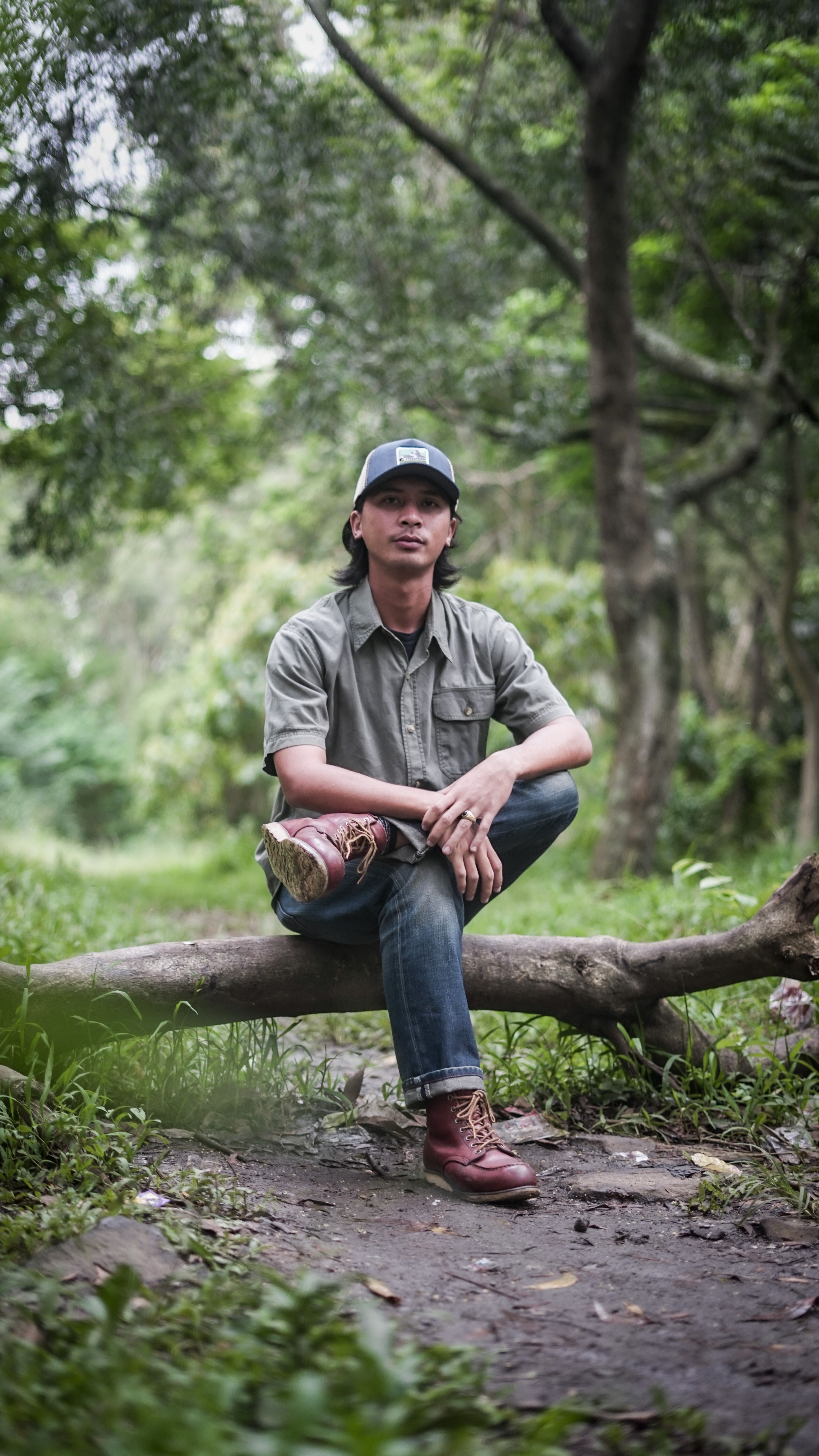 A young man in casual attire sitting on a log amidst a vibrant green forest path.