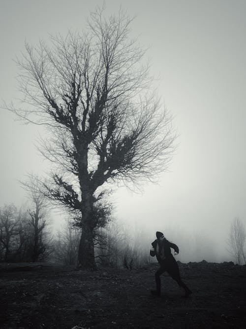 Black and White Photo of a Woman Running Next to a Spooky Tree