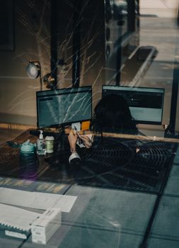Woman with black hair working at desk with dual monitors viewed through a window.