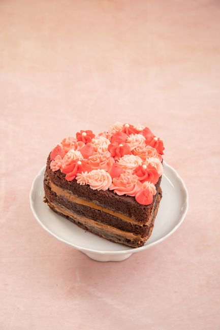 A heart-shaped chocolate cake with pink floral icing on a white pedestal against a pink background.