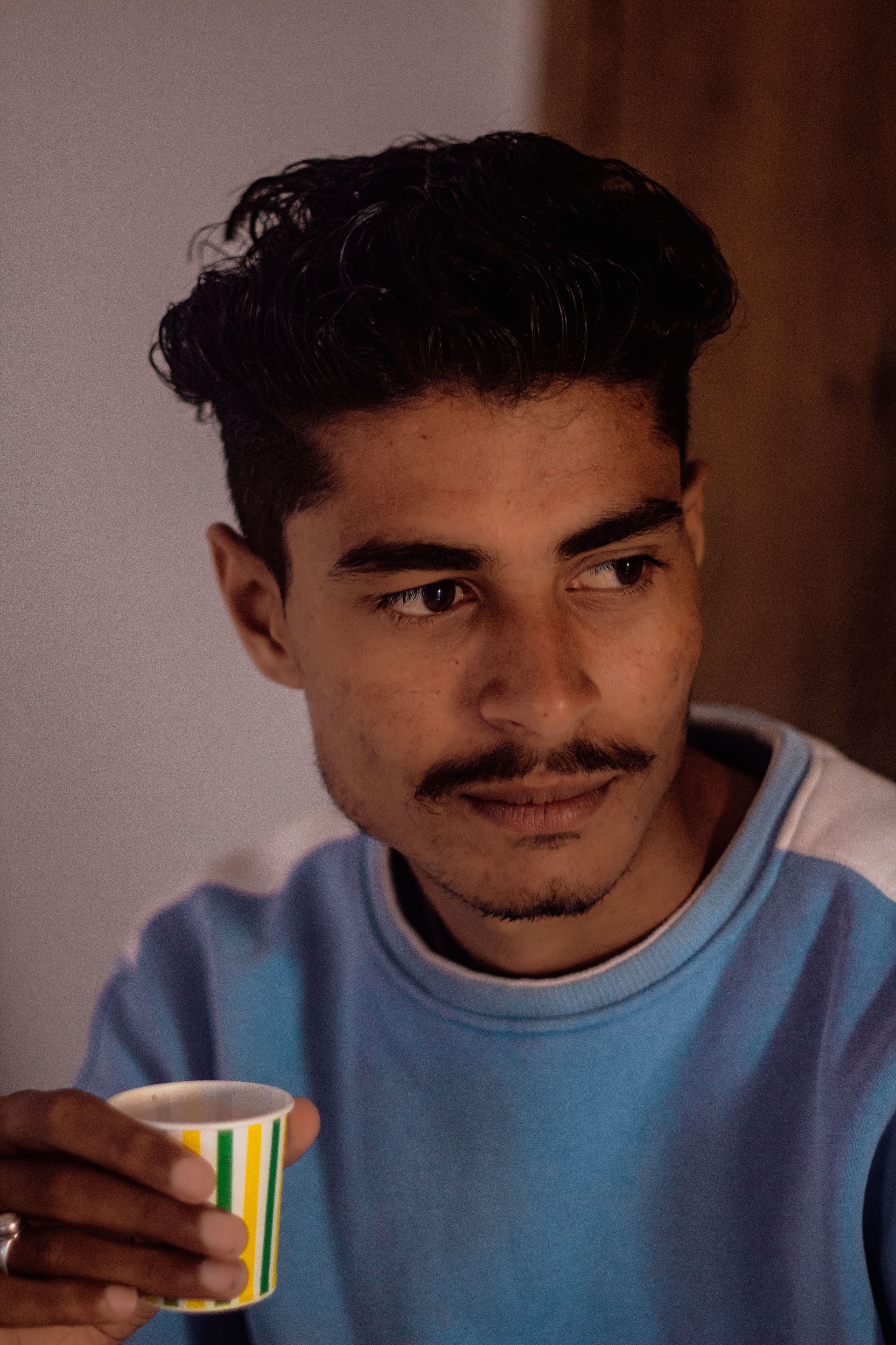 A thoughtful man with short black hair holds a cup indoors, creating a warm and introspective mood.