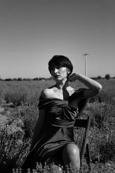 A woman in an elegant dress sits in a meadow, capturing a classic black and white portrait.